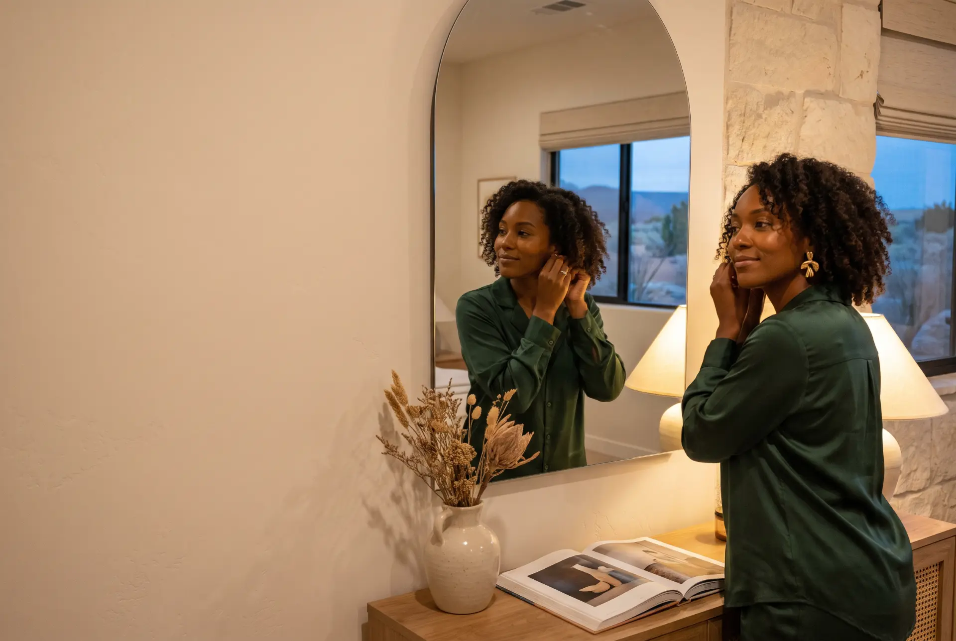 Woman putting on gold earrings in front of a mirror in a warm, softly lit home at evening.
